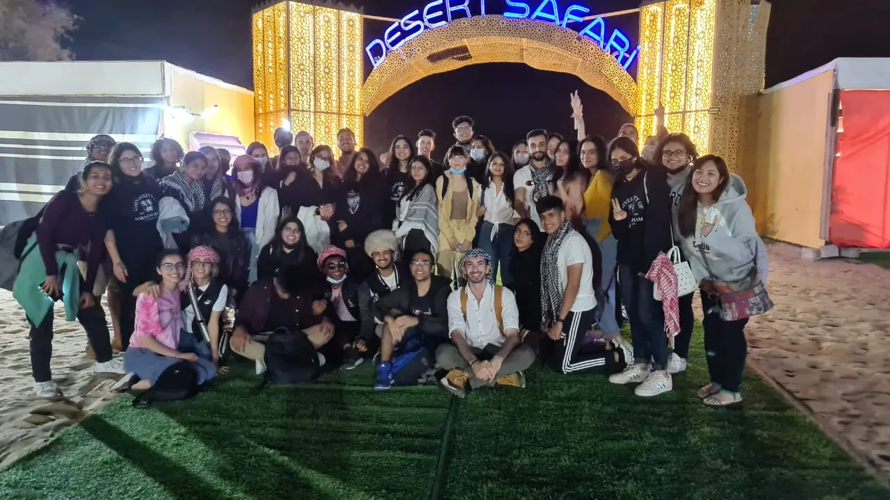 A large group of University of Birmingham Dubai students posing at night under a lit Desert Safari archway